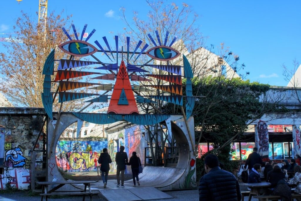 Visitors stroll under a colorful abstract entrance sculpture at the Darwin Ecosystem in Bordeaux, an alternative cultural space known for its vibrant street art and community events, a creative hub that adds a splash of modern artistic flair to the historic city.