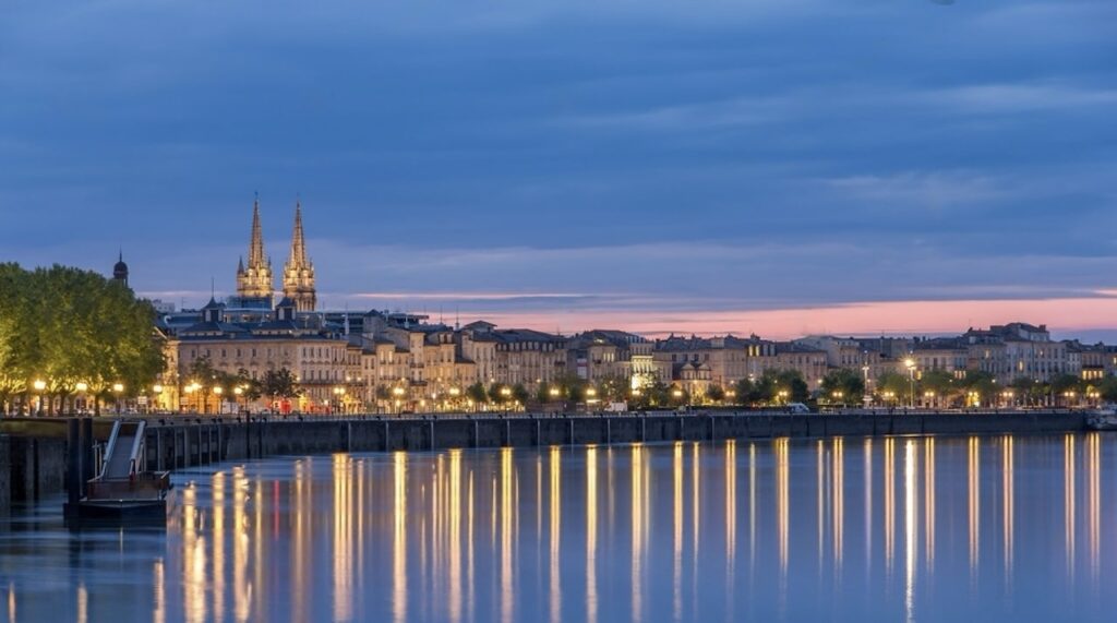 Twilight descends on the Garonne River in Bordeaux, with the city's lights and the majestic spires of the Saint-André Cathedral reflecting on the tranquil water, showcasing the city's historic grandeur and romantic ambiance, an essential experience when considering things to do in Bordeaux.
