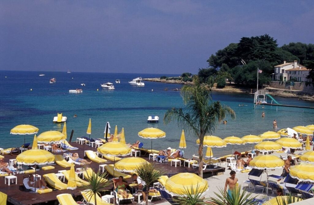 Garoupe Beach, a vibrant beach clubs in Antibes, is bustling with activity under yellow-striped umbrellas and blue lounge chairs, with bathers enjoying the clear blue waters and boats floating nearby, all under the Mediterranean sun.