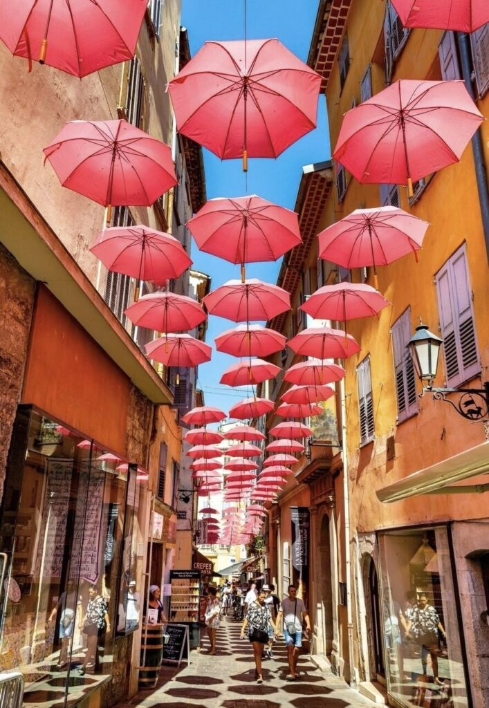 A vibrant alley in Grasse, adorned with a canopy of floating red umbrellas, presents a whimsical walkway that beckons visitors. This unique installation adds a splash of color and charm to the already picturesque setting of one of the must-visitFrench Riviera cities.