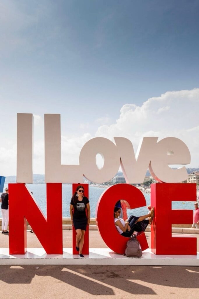 A visitor poses beside the red and white 'I love NICE' sign installation along the Promenade des Anglais, with a clear blue sky and the calm sea in the background. Perfect visual for a Nice travel guide, capturing the city's vibrant coastal charm.