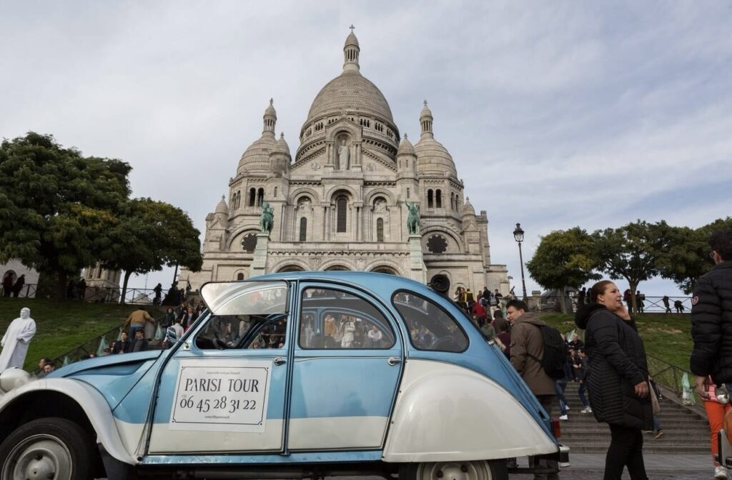 A vintage blue and white car with a 'Paris Tour' sign and contact number parked in front of the iconic Basilica of the Sacred Heart of Paris, capturing the charm of a Valentine's Day in Paris.