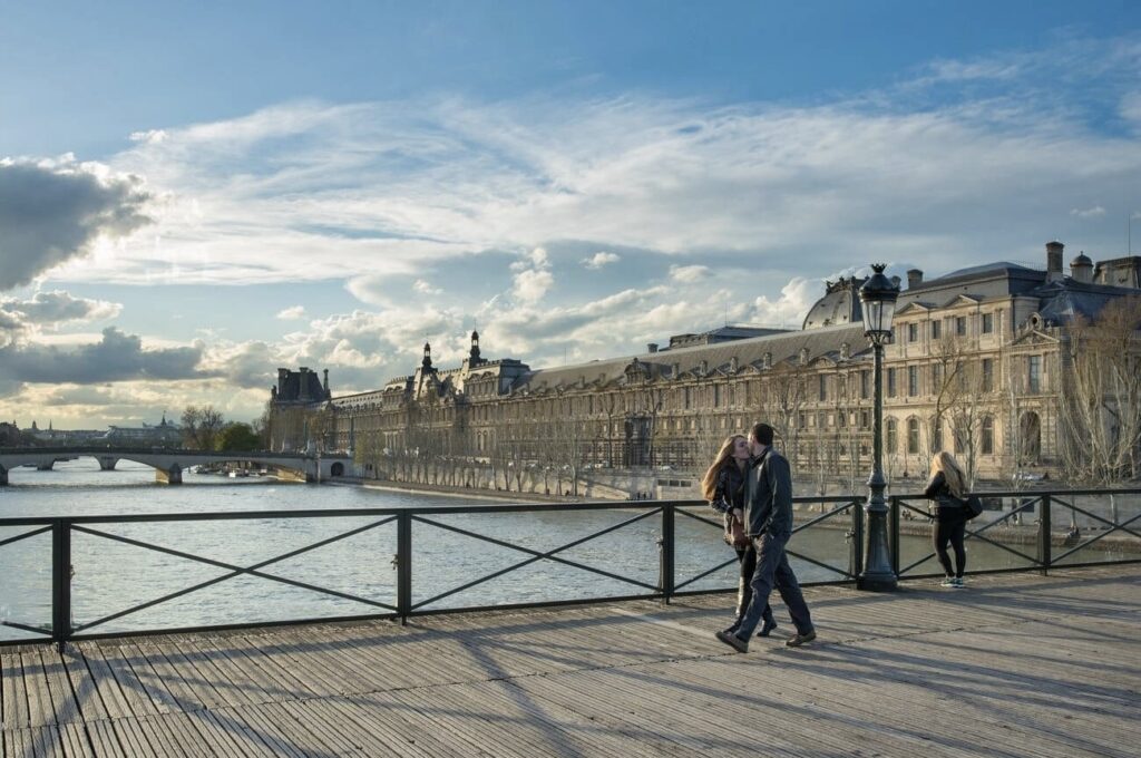 A couple shares a romantic moment on a bridge with the Louvre Museum and the Seine River in the background, encapsulating a picturesque Valentine's Day in Paris.