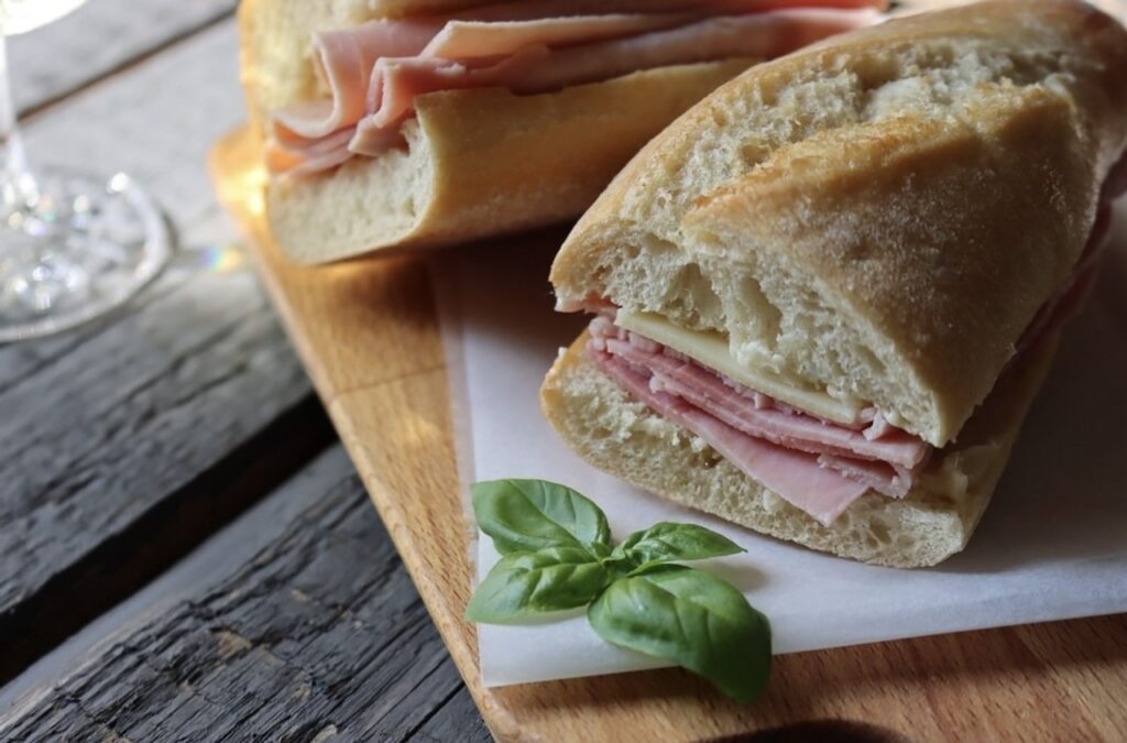 A classic Jambon-Beurre sandwich, one of the famous French foods, with layers of ham and cheese on a fresh baguette, garnished with basil leaves, presented on a wooden board with a rustic backdrop.