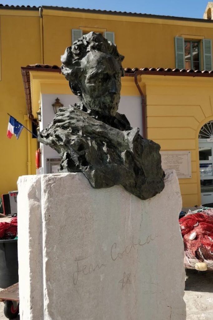Bronze statue of Jean Cocteau, poised contemplatively, set against the backdrop of a yellow building and the French flag in Villefranche-sur-Mer. The signature of Jean Cocteau is etched onto the stone pedestal, offering a cultural touchpoint for those exploring artistic things to do in Villefranche-sur-Mer.