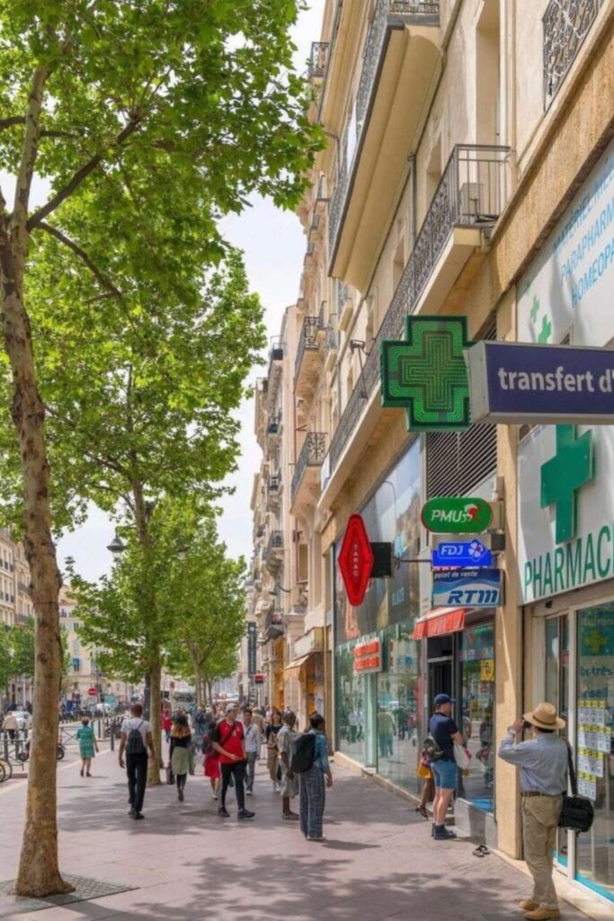 Everyday life unfolds on La Canebière, Marseille's historic high street, with pedestrians walking under green leafy trees, passing by a pharmacy and various shops, capturing the city's lively urban atmosphere as a part of the things to do in Marseille.