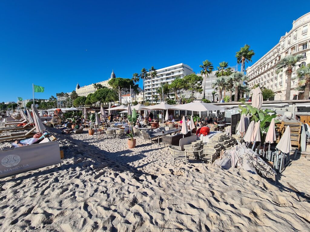 Beachgoers enjoy the sun at La Mandala, a popular Cannes beach club, with comfortable seating and umbrellas on the sand. The bustling promenade, palm trees, and elegant architecture of Cannes provide a luxurious urban beachfront atmosphere.