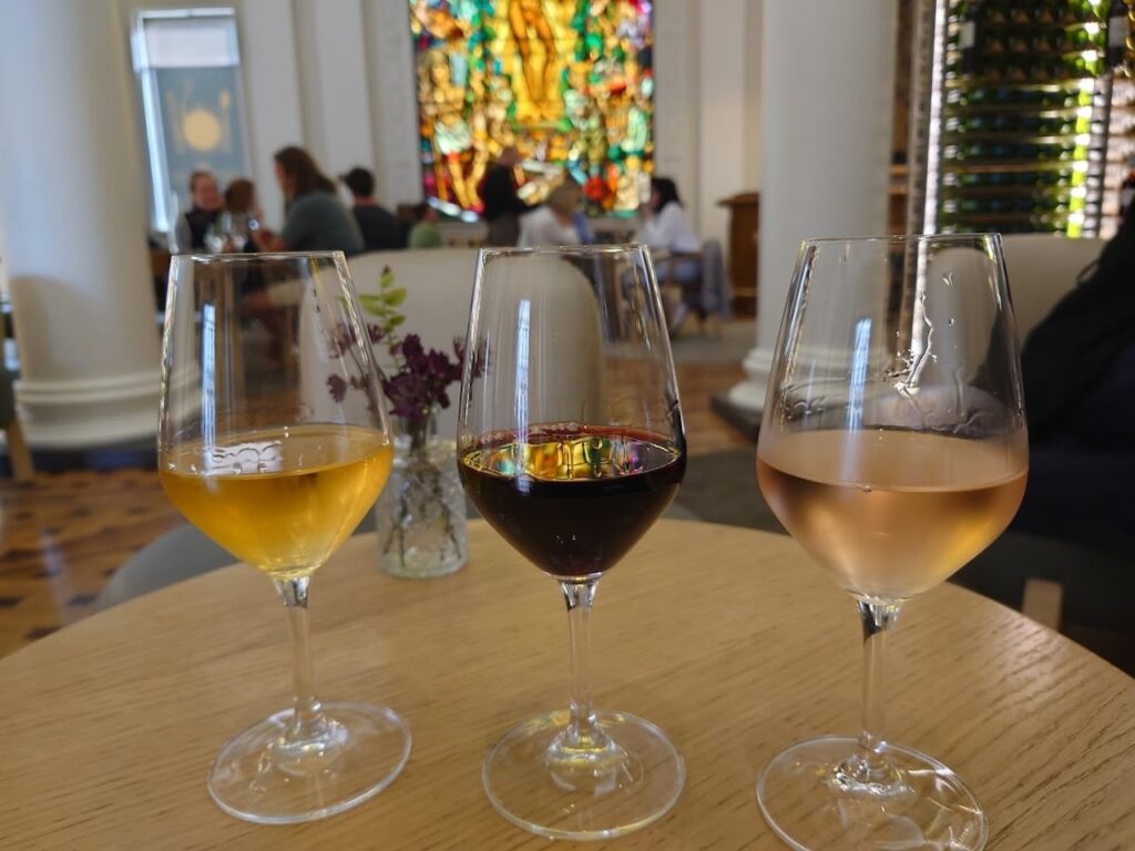 An elegant wine tasting experience at Le Bar à Vin du CIVB in Bordeaux, France, showcasing a flight of local wines with a backdrop of patrons and stained glass art. Enjoying the region's famed viniculture is one of the sophisticated things to do in Bordeaux France.