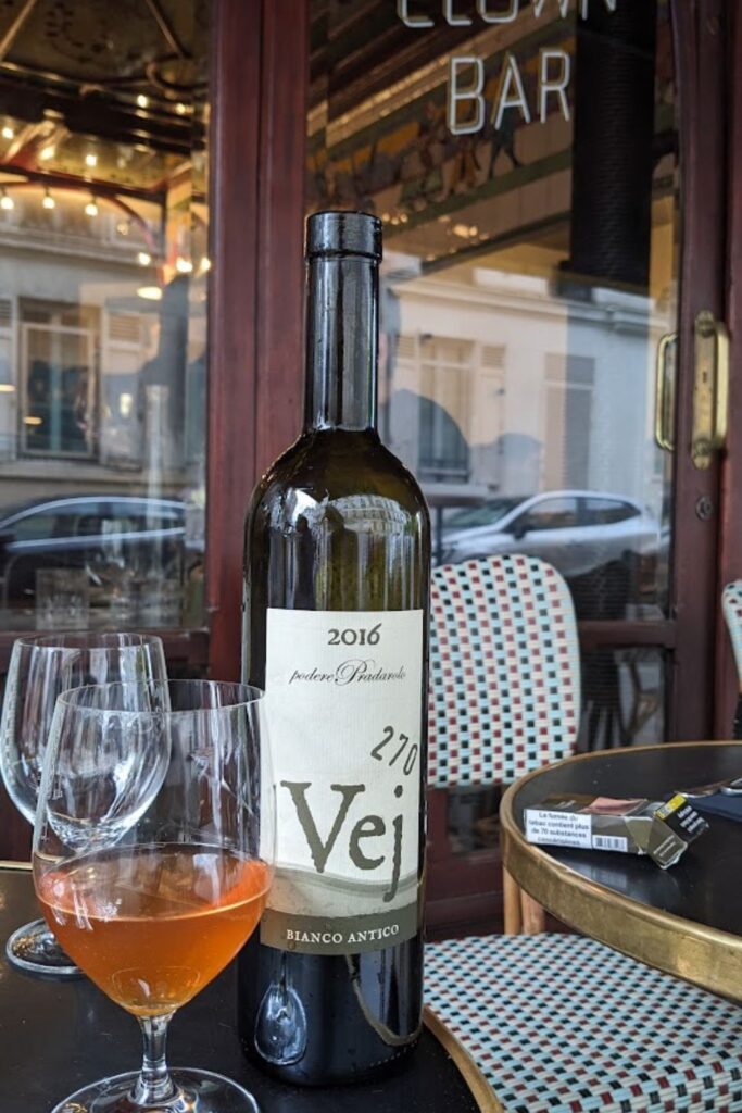 A bottle of 2016 Podere Pradarolo Vej Bianco Antico wine featured on a table with a half-filled glass, against the lively street view and iconic entrance of Le Clown Bar, a must-visit for enthusiasts exploring the best wine bars in Paris.