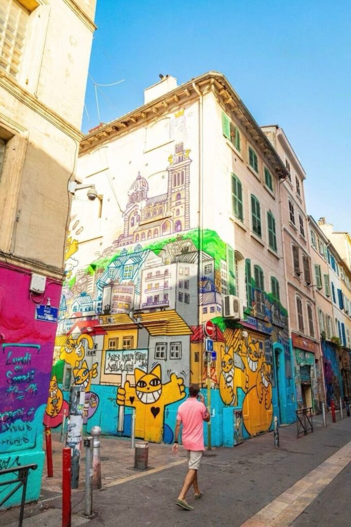 A person walks by a colorful street art mural in Le Cours Julien, Marseille, featuring vibrant caricatures and urban architecture drawings, reflecting the city's lively street art scene as one of the unique things to do in Marseille.