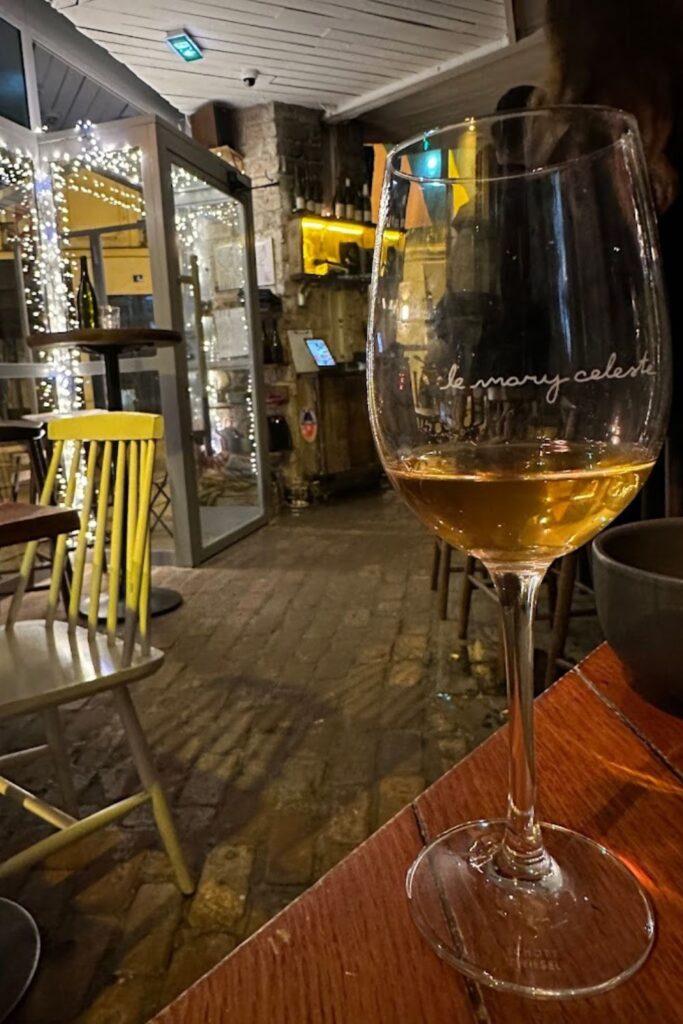 A glass of golden wine rests on a table with the inscription 'Le Mary Celeste,' offering a glimpse into the cozy, festive atmosphere of Paris's revered wine bars, complete with twinkling lights and classic stone pavement views.