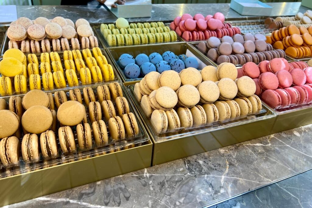 An assortment of colorful macarons, iconic of famous French foods, displayed in neat rows with a variety of flavors such as lemon, pistachio, and raspberry, showcased on a marble countertop invitingly for sweet indulgence.