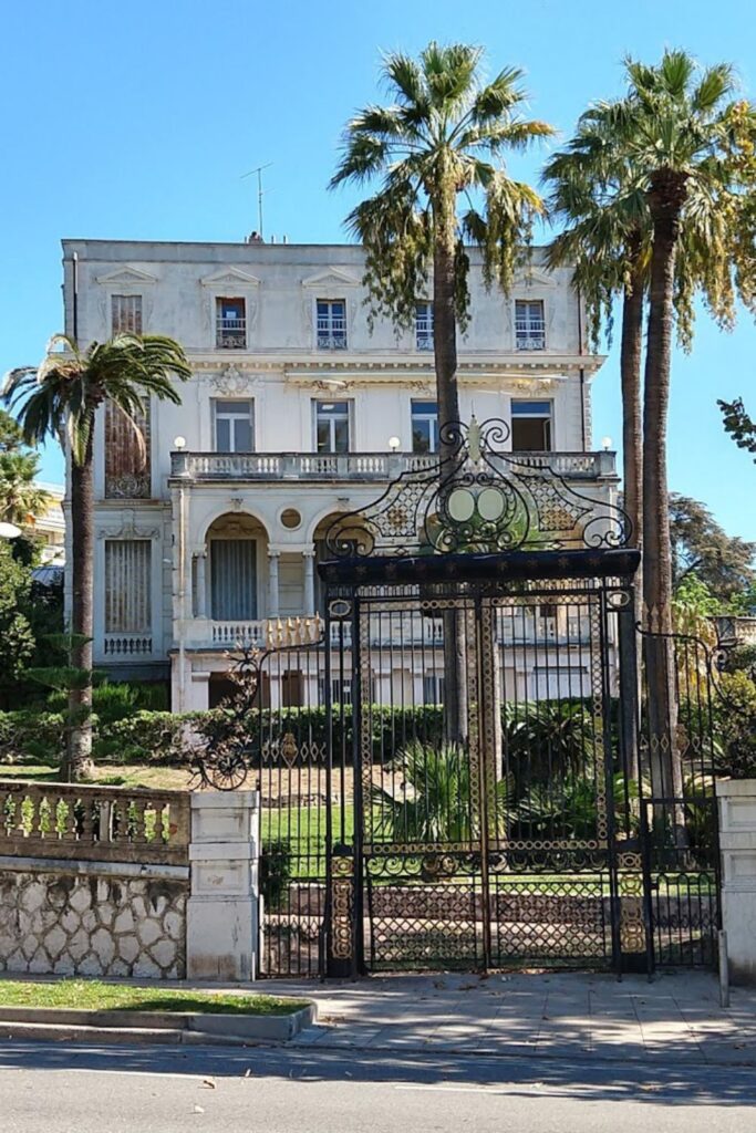 Elegant wrought-iron gates open to a grand, vintage villa in Nice, flanked by tall palm trees against a clear blue sky. This image encapsulates the architectural beauty and historical residences that add to the charm of Nice, perfect for a travel guide exploring the city's hidden gems.