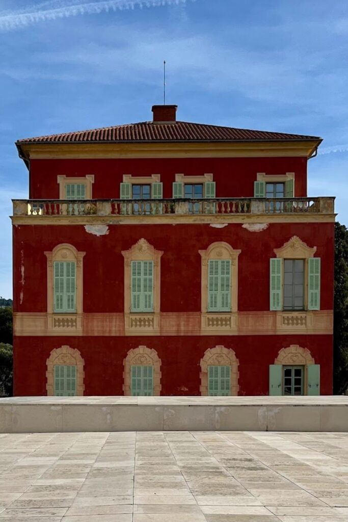 The Matisse Museum in Nice, a radiant red ochre building with classic French windows and a terracotta roof, standing majestically under a blue sky. Its striking facade is a focal point in a Nice travel guide, inviting visitors to explore the artistic treasures within.