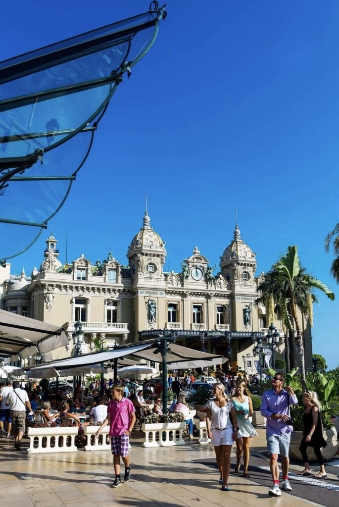 Alt text: "The bustling atmosphere outside the grand Casino de Monte-Carlo, with visitors strolling and enjoying the sunny day. Framed by a brilliant blue sky and tall palms, this iconic casino is a jewel among the must-visit French Riviera cities.