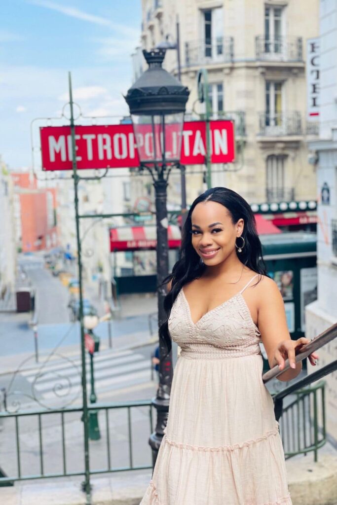 A beaming woman in a flowing beige dress stands before the iconic red METROPOLITAIN sign of the Parisian subway in Montmartre, capturing a charming moment at one of the best Paris instagram spots