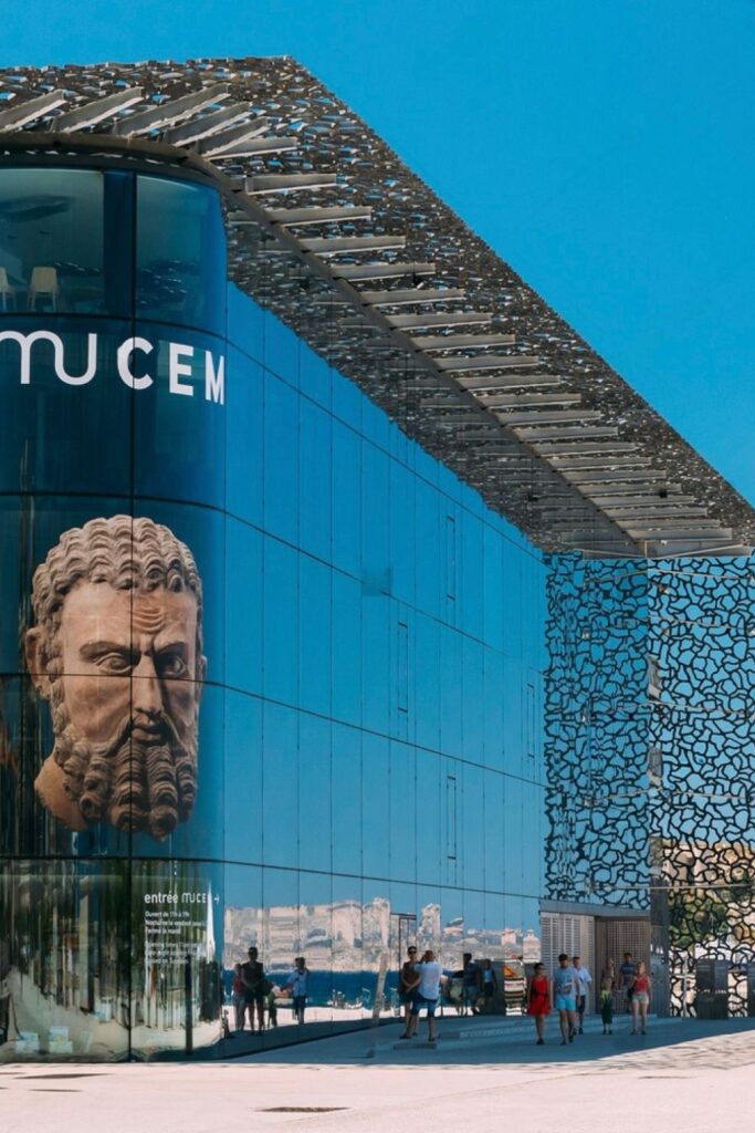 Visitors approach the striking, modern facade of MuCEM (Museum of European and Mediterranean Civilisations) in Marseille, with a large face sculpture reflected on the glass and an intricate concrete lattice, symbolizing cultural exploration among the things to do in Marseille.