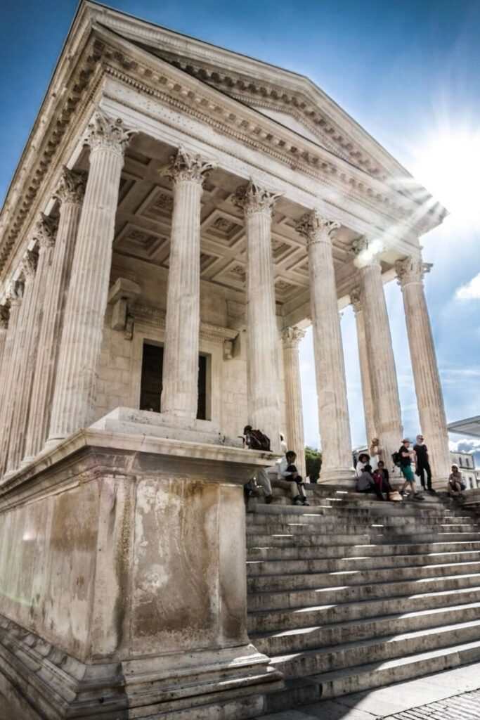 Visitors gather on the steps of the grand Maison Carrée in Nîmes, France, an impeccably preserved Roman temple basking in the sunlight, a striking historical highlight for a day trips from Montpellier