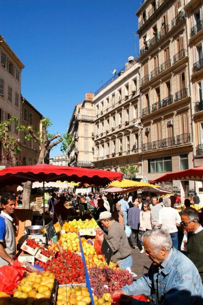 Bustling scene at the Noailles Market in Marseille, with locals and visitors browsing through colorful stalls of fresh fruits under the shade of red and yellow canopies, nestled among traditional French architecture, a lively experience for food enthusiasts exploring things to do in Marseille.
