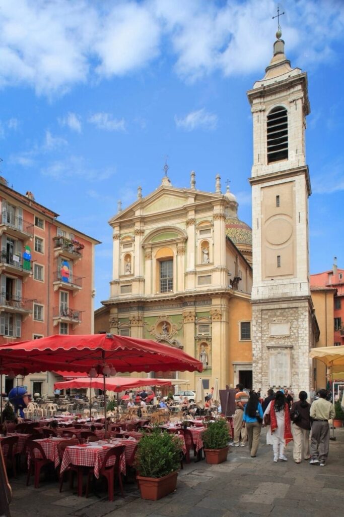 Outdoor café scene in the Old Town of Nice, with red-checkered tablecloths and a backdrop of charming pastel-colored buildings and the Baroque façade of the Cathedral Sainte-Réparate. A picturesque spot that captures the essence of Nice's vibrant dining culture, perfect for a travel guide.