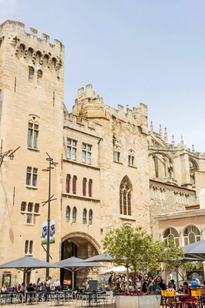 The bustling square in front of the Palace of the Archbishops in Narbonne, with outdoor cafés and relaxed patrons, offers a blend of historical architecture and modern leisure, ideal for day trips from Montpellier.