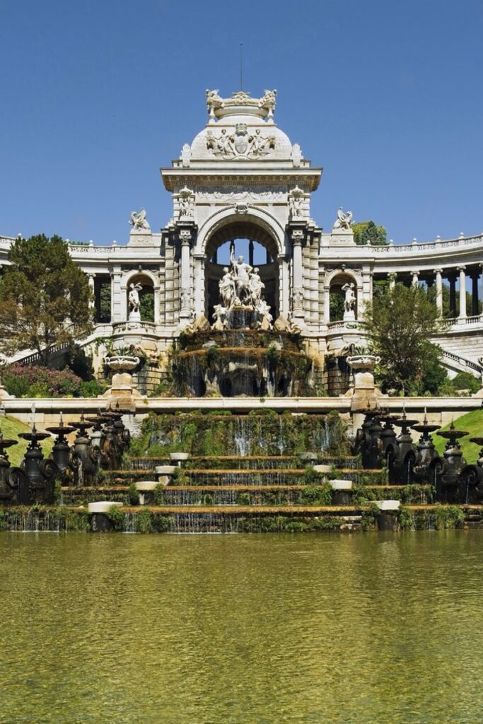 The majestic Palais Longchamp in Marseille, with its intricate stone fountain and cascading water, surrounded by lush greenery and reflected in a tranquil pond, a historical site that is a must-see for those interested in the architectural things to do in Marseille.