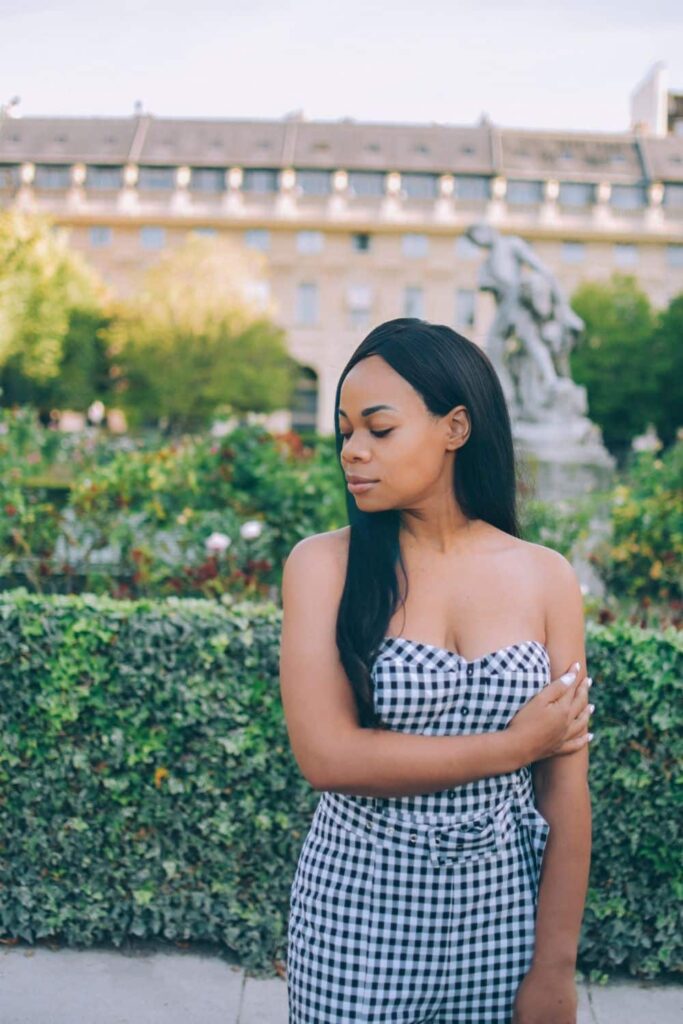A serene woman in a gingham dress closes her eyes and enjoys a tranquil moment in the Palais-Royal Garden, with lush greenery and an elegant statue in the background, a hidden gem among the best Paris Instagram spots.