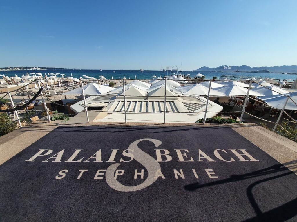The entrance mat to Palais Beach Stephanie with its logo prominently displayed, leading to a panoramic view of a Cannes beach club, showcasing rows of white sunbeds and umbrellas, with the serene Mediterranean Sea and distant mountains completing this idyllic coastal setting.