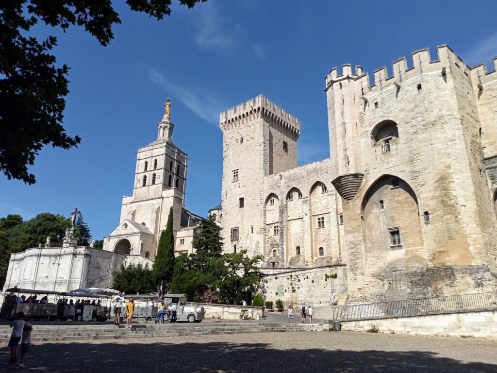 The majestic Palais des Papes in Avignon, with its fortified stone walls and Gothic architecture, under a clear blue sky framed by greenery, a historical landmark that's a staple on the list of the best things to do near Arles.