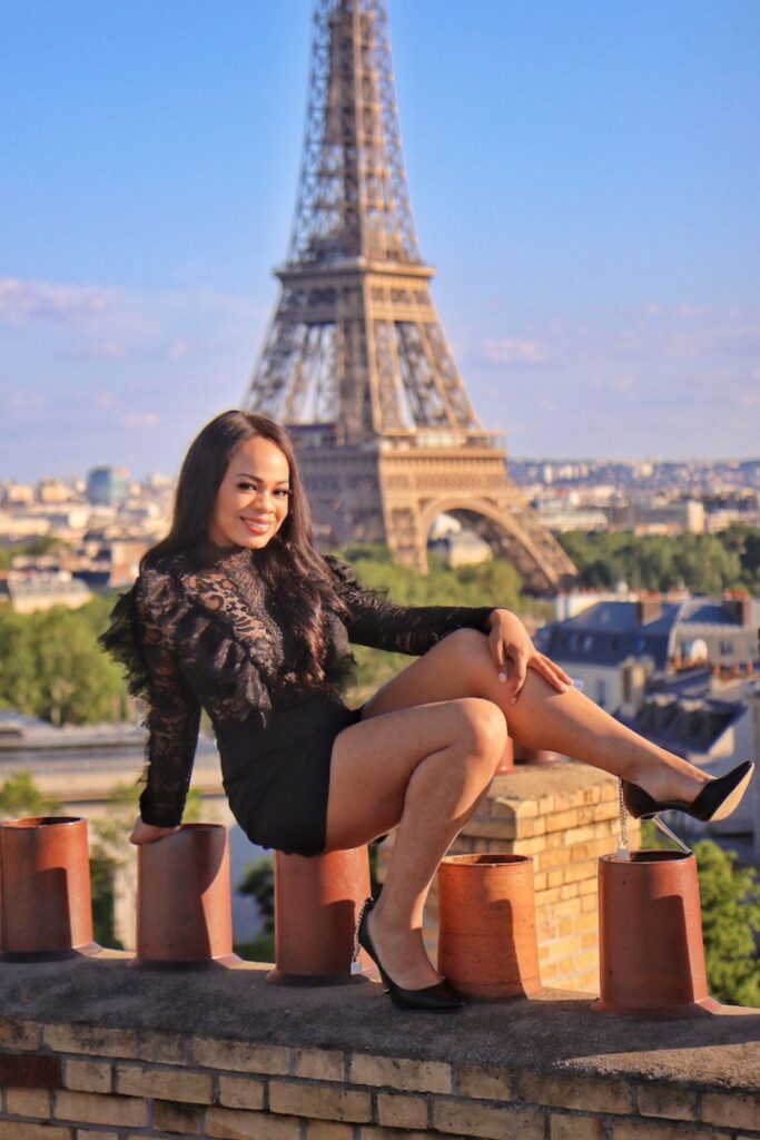 A woman dressed in elegant black lace poses seated on a rooftop ledge, with a stunning view of the Eiffel Tower and Parisian rooftops in the background, epitomizing the allure of the best Paris instagram spots