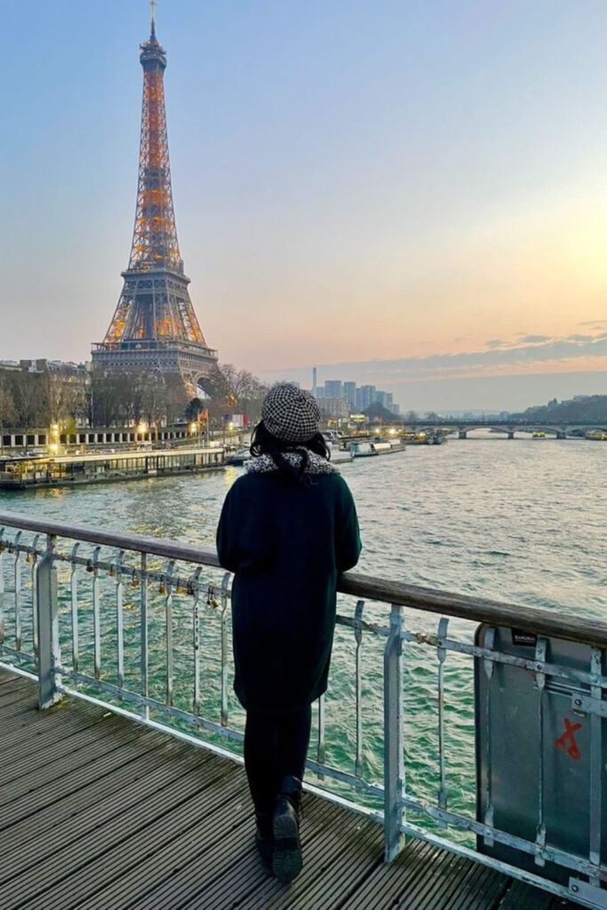 A woman stands on the Passerelle Debilly, gazing at the illuminated Eiffel Tower during twilight. This serene vantage point is a less-known but stunning Paris instagram spots, perfect for reflective moments.