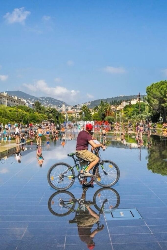 A cyclist pauses on the reflective surface of the Miroir d'eau fountain at Place Masséna in Nice, with the city's hills in the distance and people enjoying the water features. A must-visit attraction for a Nice travel guide, showing a slice of daily life in this dynamic city.