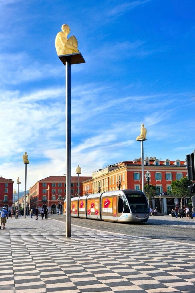 Place Masséna, the vibrant heart of Nice, with the modern tramway gliding past the iconic checkered pavement and striking stone sculptures atop poles. This bustling square is a key highlight for any Nice travel guide, illustrating the city's fusion of historic charm and contemporary living.