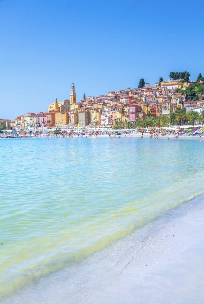 The crystal-clear waters of Plage des Sablettes in Menton gently kiss the shore, with the city’s pastel-colored buildings cascading up the hillside under a brilliant blue sky. This picturesque beachfront is a highlight in Menton, epitomizing the allure of must-visit French Riviera cities.