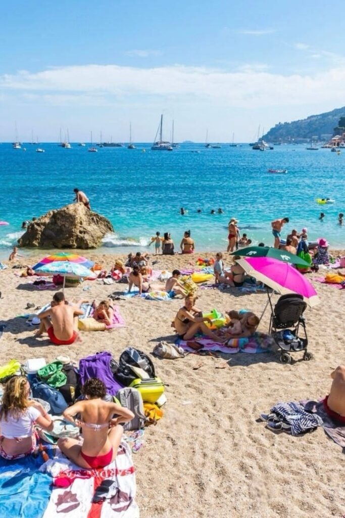 Bustling summer day at Plage des Marinières in Villefranche-sur-Mer, with people sunbathing, swimming, and enjoying the clear blue waters and sailboats in the distance. The beach scene, complete with colorful umbrellas and the hillside town backdrop, exemplifies relaxing activities and things to do in Villefranche-sur-Mer.