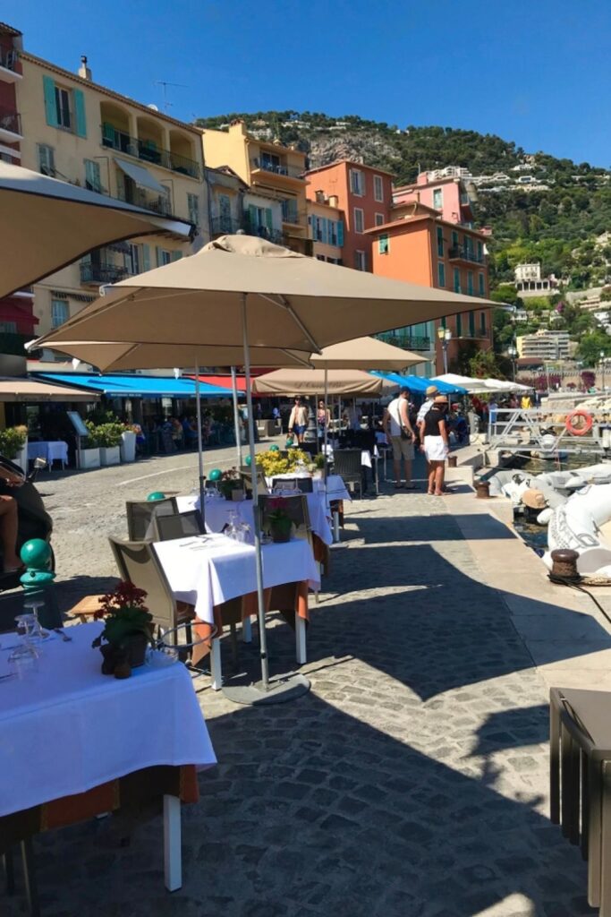 Alfresco dining on Quai de l'Amiral Courbet in Villefranche-sur-Mer, where elegantly set tables under umbrellas line the vibrant waterfront, with colorful facades and hillside views in the background. This charming setting offers a perfect culinary experience for visitors searching for things to do in Villefranche-sur-Mer.