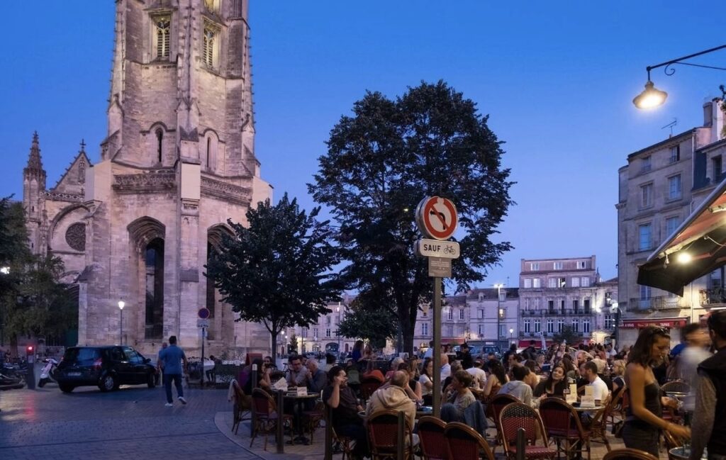 A bustling evening scene at a street café in Bordeaux, with patrons enjoying meals under the ambient glow of streetlights, beside an ornate church and a 'no entry except bicycles' traffic sign, encapsulating the vibrant nightlife and architectural beauty as one of the things to do in Bordeaux.