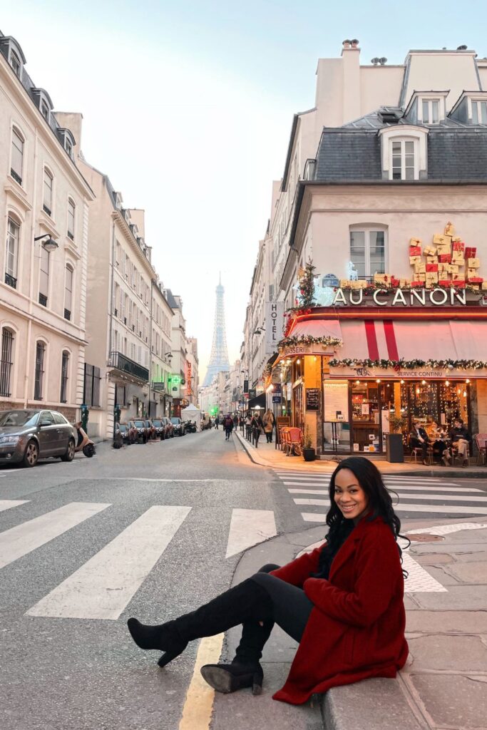 Seated on the curb of Rue Saint-Dominique, a woman in a vibrant red coat and knee-high boots flashes a charming smile, with the Eiffel Tower creating an iconic backdrop, a quintessential Paris instagram spots.