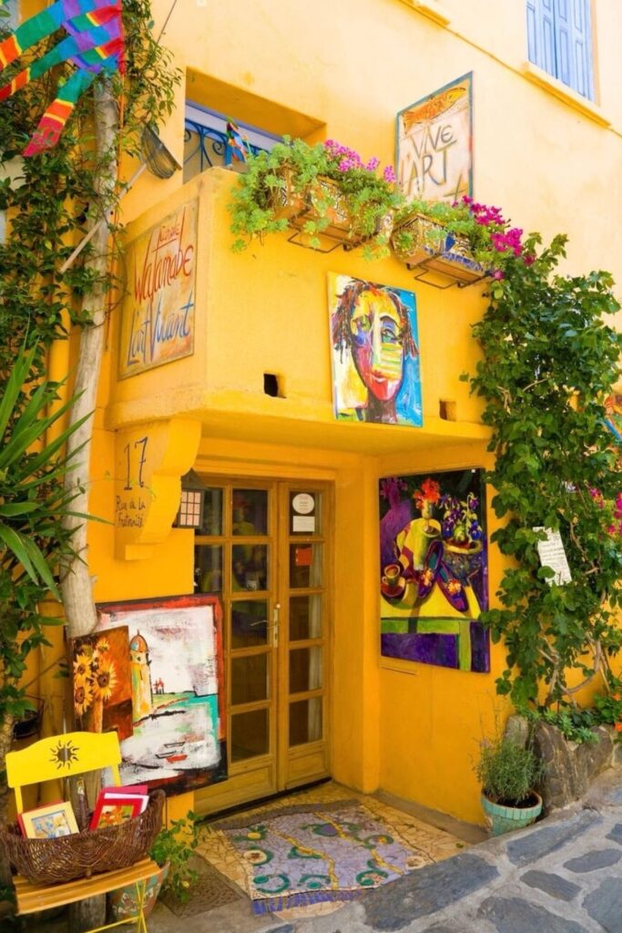 The vibrant art-filled entrance to 'Madame Cotlliure' on Rue de la Fraternité in Collioure, France, invites onlookers with its warm yellow walls and colorful artwork, a charming find on a day trips from Montpellier.