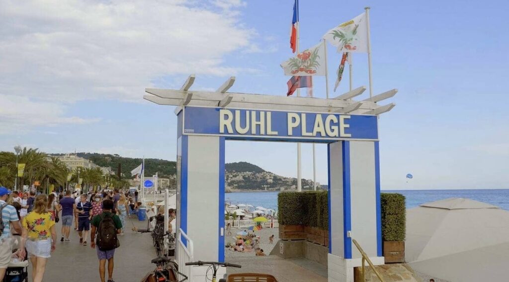 Entrance to Ruhl Plage, one of the best Beach Clubs in Nice, framed by a white and blue archway with fluttering flags above, inviting visitors down a lively promenade towards the sun-drenched Mediterranean beachfront.