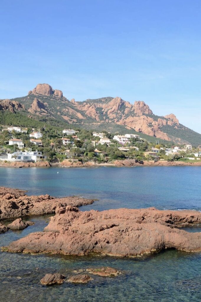 Alt text: "The tranquil cove of Saint-Raphaël, with its crystal-clear waters and distinctive red rocks, lies before a backdrop of rugged hills dotted with Mediterranean villas. This scenic landscape is a highlight of Saint-Raphaël, a gem among the must-visit French Riviera cities.