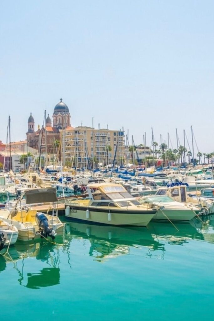 Alt text: "The serene marina of Saint-Raphaël is a hive of nautical activity, with a variety of boats moored in the calm turquoise waters. Dominated by the striking architecture of the town's church in the background, Saint-Raphaël is a coastal haven that stands out among the must-visit French Riviera cities.