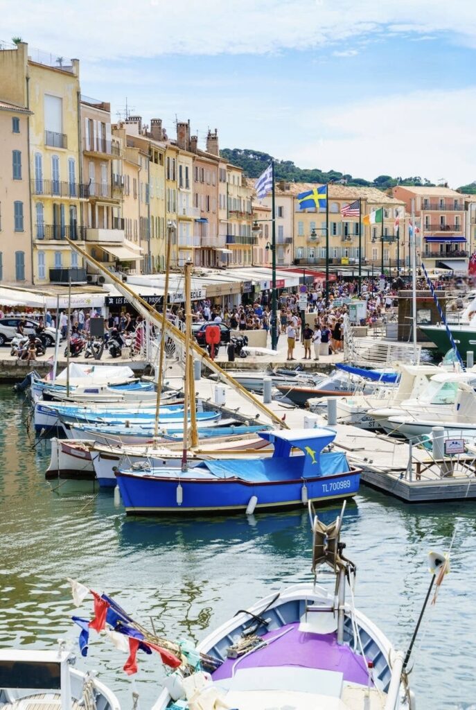 Alt text: "A lively marina scene at Saint-Tropez, with traditional boats moored in clear waters and crowds of people exploring the waterfront promenade. The colorful facades of buildings reflect the town's vibrant character, a staple of the must-visit French Riviera cities.