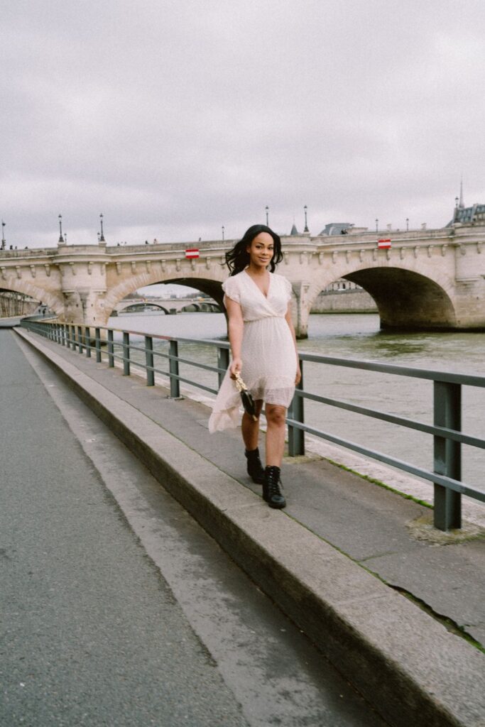 A woman in a delicate white dress and combat boots poses with the Seine River behind her, highlighting the juxtaposition of romantic and edgy styles at this quintessential Paris instagram spots, with the Pont Neuf in the distance.