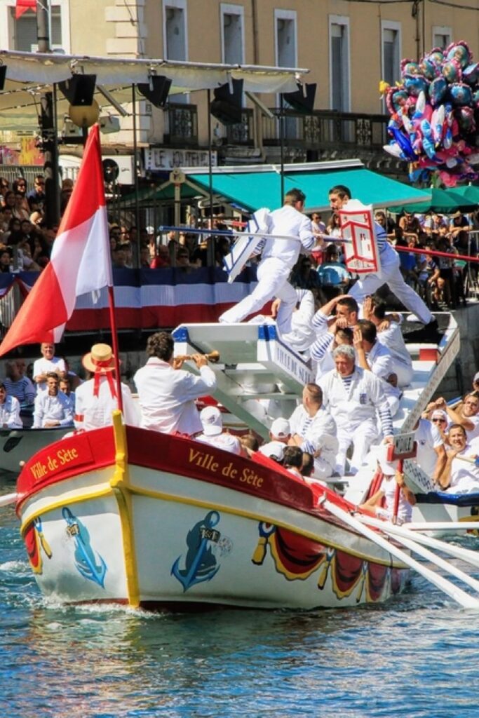 Enthusiastic participants engage in traditional water jousting in Sète, France, with onlookers cheering from the decorated boats and riverside. This lively cultural event is a unique spectacle to enjoy on a day trips from Montpellier