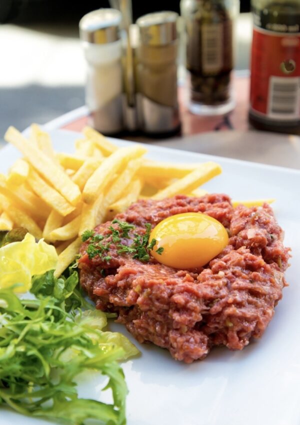 Savoring famous French foods, this image shows a delectable Steak Tartare with a vibrant raw egg yolk on top, paired with golden French fries and a light green salad, served on a sunny café table with condiments in the background.