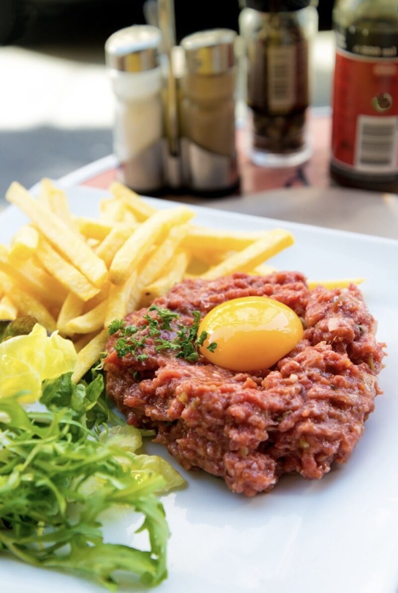 Savoring famous French foods, this image shows a delectable Steak Tartare with a vibrant raw egg yolk on top, paired with golden French fries and a light green salad, served on a sunny café table with condiments in the background.