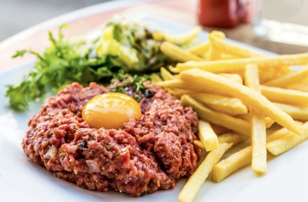 A traditional Steak Tartare, an iconic item among famous French foods, topped with a raw egg yolk and served with a side of crisp French fries and a delicate green salad, embodying the bold flavors of France.