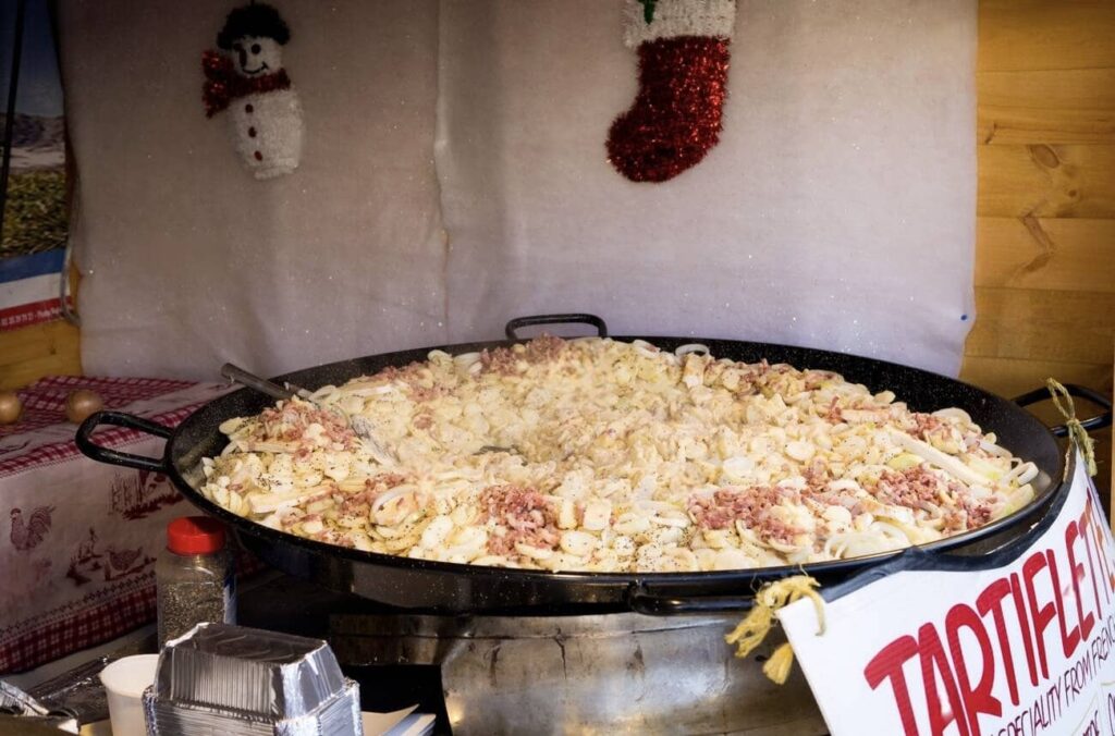 A large, steaming pan of Tartiflette, a famous French food, with golden potatoes, luscious melted cheese, and bits of bacon, ready to be served, with a festive background featuring a snowman and stocking decorations.