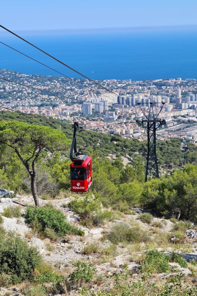 The red Toulon cable car ascends against a stunning vista of the cityscape and Mediterranean Sea, highlighting the adventurous side of Toulon, a must-visit French Riviera cities. The lush greenery and rocky terrain below add to the scenic journey up Mount Faron.
